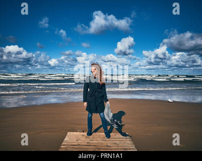 Jeune fille rousse vêtue d'un manteau noir promenades le long du littoral. Un fort vent a attisé les boucles de la jeune fille. Banque D'Images