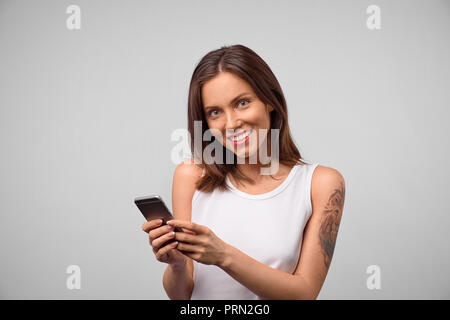 Brunette souriante jeune femme montrant des dents blanches en utilisant un téléphone cellulaire, messagerie instantanée, d'être heureux au texte avec son petit ami. Les technologies modernes et co Banque D'Images