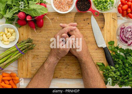 Man resting hands on planche à découper en bois bordée par de multiples légumes y compris le poivron vert et les carottes à côté des petits bols d'épices Banque D'Images