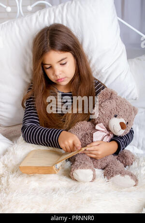 Petite fille couchée sur le lit avec son ours en peluche préféré et la lecture d'un livre avant le coucher Banque D'Images