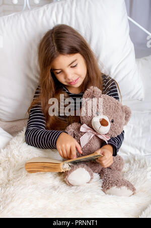 Petite fille couchée sur le lit avec son ours en peluche préféré et la lecture d'un livre avant le coucher Banque D'Images