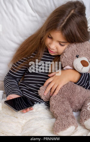 Petite fille couchée sur le lit avec son ours en peluche préféré et jouer un smartphone Banque D'Images