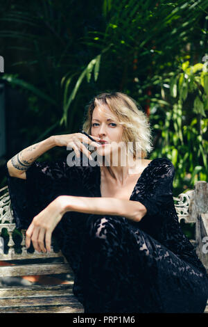 Femme blonde en vêtements noirs cigarette alors qu'elle repose sur le banc sur terrasse, Ubud, Bali, Indonésie Banque D'Images