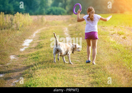 Une jeune fille avec un chien court le long d'une route de campagne dans un champ en été. La fille tient dans sa main un anneau pour la formation Banque D'Images