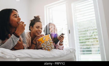 Trois jeunes filles lying on bed eating popcorns à partir d'un bain à remous vous regardez la télévision. Pour profiter de regarder la télévision à la maison pendant une soirée pyjama. Banque D'Images