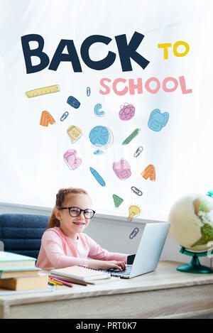 Adorable gamin dans eyeglasses smiling at camera while étudie avec ordinateur portable à la maison avec des icônes et 'lettrage' retour à l'école Banque D'Images