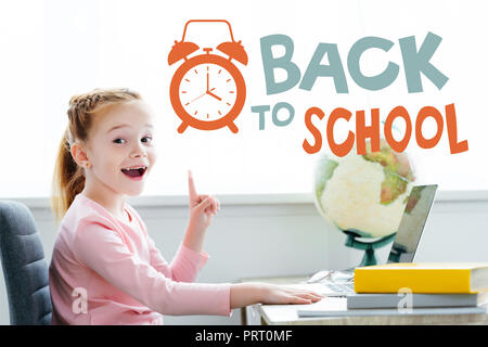 Cheerful redhead enfant pointe vers le haut avec le doigt et smiling at camera tout en étudiant avec des livres et l'ordinateur portable à la maison avec "retour à l'école' avec lettrage Banque D'Images