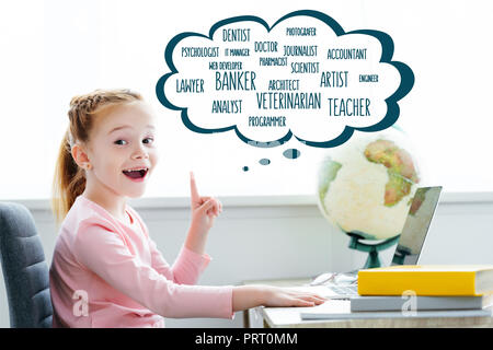 Cheerful redhead enfant qui étudie avec des livres et d'ordinateurs portables et pointant vers le haut sur les mots de différentes professions en bulle Banque D'Images