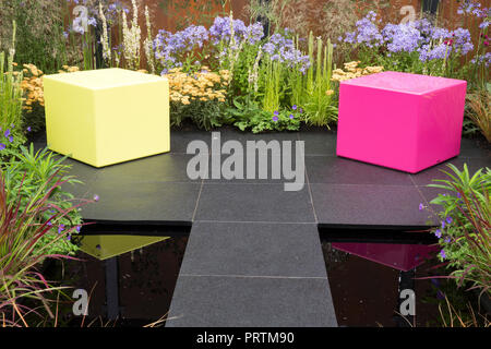 Moderne Pink et jaune boîte en plastique cube siège sombre moderne pierre pavée patio zone petit étang réfléchissant foncé coloré bordures de fleurs mélangées Banque D'Images