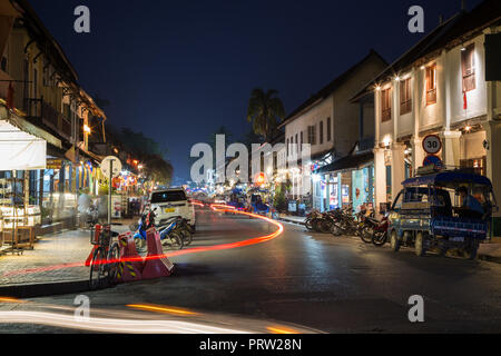 Des scooters, des voitures et des taxis à trois roues (jumbo ou tuk-tuk), plusieurs personnes, allumé les bâtiments et les voitures de l'époque coloniale' light trails in Luang Prabang au crépuscule. Banque D'Images