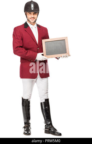 Jeune cavalier souriant en uniforme holding blank chalkboard and looking at camera isolated on white Banque D'Images