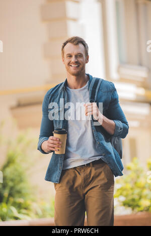 Heureux jeune homme avec du café pour aller marcher sur la rue et un sac à dos et looking at camera Banque D'Images