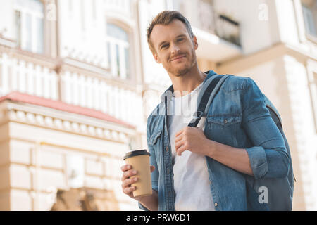 Vue de dessous du beau jeune homme avec du café pour aller marcher sur la rue et un sac à dos et looking at camera Banque D'Images