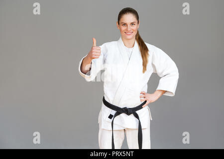 Jeune femme sportive positive en kimono blanc et noir permanent de la courroie, Thumbs up, looking at camera avec sourire à pleines dents. Concept arts martiaux japonais. Banque D'Images