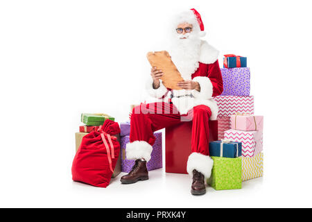 Père Noël en lecture lunettes vieux parchemin et assis sur des tas de boîtes cadeaux isolé sur fond blanc Banque D'Images