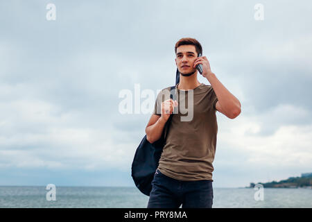 Jeune homme souriant avec guitare acoustique marche sur nuageux beach talking on phone Banque D'Images