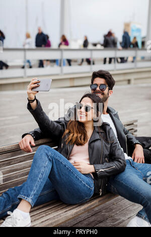 Espagne, Barcelone, happy young couple reposant sur un banc en tenant un selfies Banque D'Images