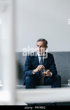 Mature businessman avec verre de café assis sur le canapé dans son bureau à la fenêtre de Banque D'Images