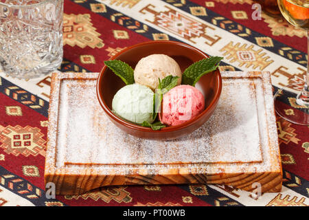 La crème glacée avec des feuilles de menthe servi dans un bol sur le dessus d'une planche de bois sur une nappe traditionnel arménien. 3 boules ; framboise, pistache et Caramel Banque D'Images