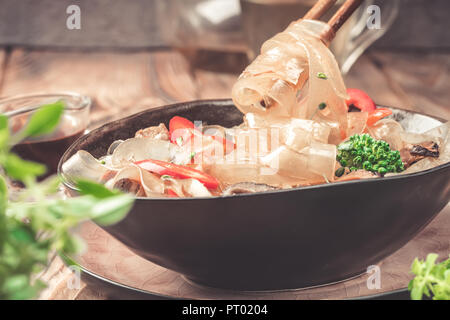 Prendre la main avec des baguettes asiatiques chaudes d'épaisseur de verre plat épicé couper les nouilles à la sauce aigre-douce haricots viande légumes brocoli poivron poireau dans bol sur une table de cuisine sauce soja et thé vert en électrique et mug. Banque D'Images