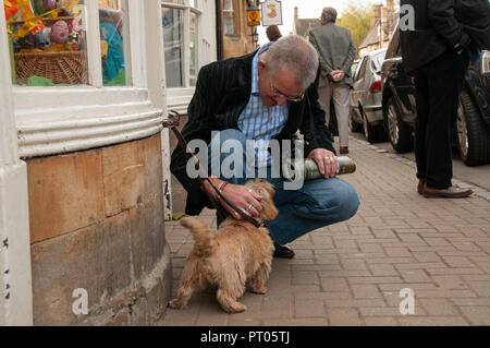 Plus l'homme habillés en chemise et blazer, tenant un magazine roulé vers le bas pour s'accroupir un peu avc terrier attaché sur le trottoir à l'extérieur. Banque D'Images