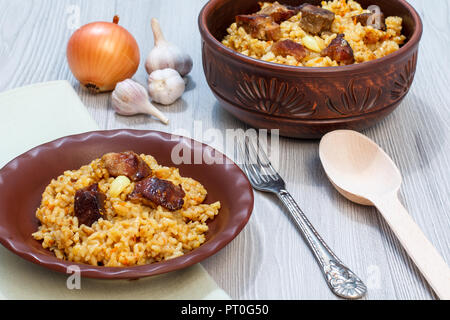 Riz pilaf sur plaque d'argile et le bol avec la viande et les légumes - l'oignon, l'ail, le poivron rouge, le poivron. Cuillère en bois et en métal à côté de la fourche vaisselle Banque D'Images