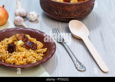 Riz pilaf sur plaque d'argile et le bol avec la viande et les légumes - l'oignon, l'ail, le poivron rouge, le poivron. Cuillère en bois et en métal à côté de la fourche vaisselle Banque D'Images