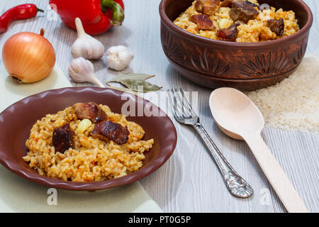 Riz pilaf sur plaque d'argile et le bol avec la viande et les légumes - l'oignon, l'ail, le poivron rouge, le poivron. Cuillère en bois et en métal à côté de la fourche vaisselle Banque D'Images