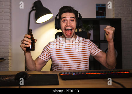 Portrait de gamer joyeux guy portant des écouteurs en criant et boire de la bière alors qu'il était assis au bureau avec ordinateur dans la pièce Banque D'Images