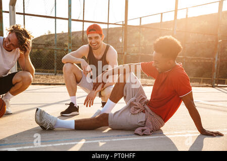 Groupe de jeunes joueurs de basket-ball hommes multiethnique gaie repose au sol, parler de sport Banque D'Images