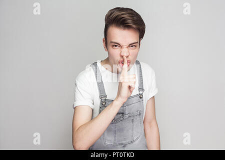 Portrait de jeune homme en colère brune dans un style décontracté avec t-shirt blanc et une salopette en jean et permanent à la caméra et au calme. montrant des signes d'indo Banque D'Images
