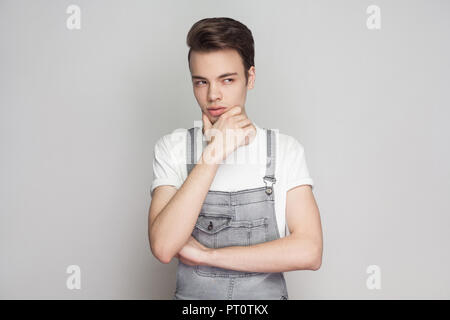 Portrait of young brunette homme en style casual avec t-shirt blanc et une salopette en jean et permanent à l'écart, de toucher le menton et la pensée. Banque D'Images