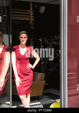 Femme portant robe bourgogne debout à la fenêtre dans sunshine Banque D'Images