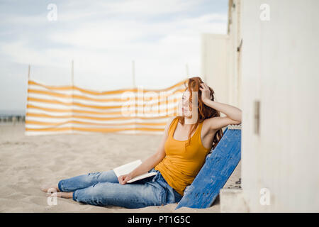 Redheaded femme assise en face de cabine de plage, la lecture d'un livre Banque D'Images