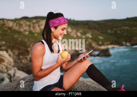Sportive woman sitting on rocks dans la soirée, la tenue et l'utilisation de smartphone Blackberry apple Banque D'Images