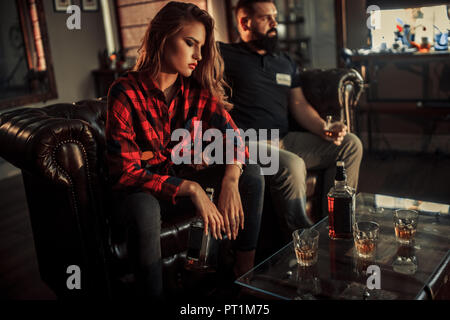 Jeune couple est assis et reposant sur un canapé dans la salle avec une bouteille et des verres de whisky. Banque D'Images