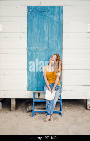 Redheaded femme assise en face de cabine de plage, la lecture d'un livre Banque D'Images