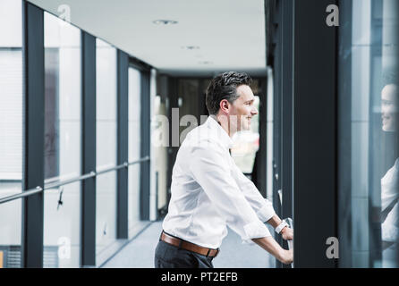 Smiling businessman looking out of window in office corridor Banque D'Images