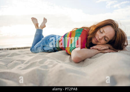 Femme rousse allongée dans le sable sur la plage, les yeux fermés Banque D'Images