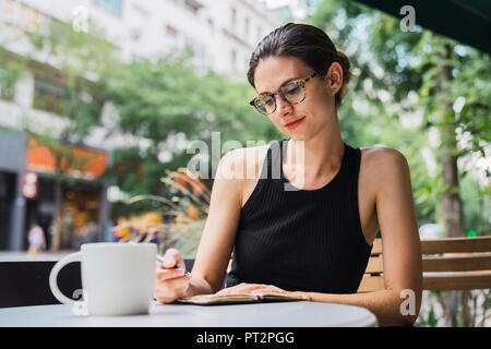 Jeune femme assise dans un café, le remue-méninges, la rédaction de notes dans l'ordinateur portable Banque D'Images