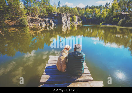 Un homme avec un Labrador retriever dog est assis sur une terrasse en bois sur une belle côte rocheuse d'un lac et s'intéresse à l'eau Banque D'Images