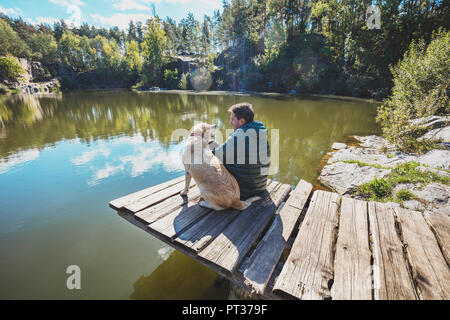 Un homme avec un Labrador retriever dog est assis sur une terrasse en bois sur une belle côte rocheuse d'un lac et s'intéresse à l'eau Banque D'Images