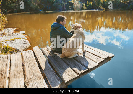 Un homme avec un Labrador retriever dog est assis sur une terrasse en bois sur une belle côte rocheuse d'un lac et s'intéresse à l'eau Banque D'Images