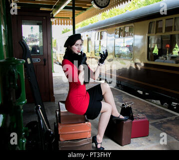 Vintage belle femme portant robe rouge et béret noir, assis sur des valises, à la recherche de miroir et d'appliquer son rouge à lèvres. Banque D'Images