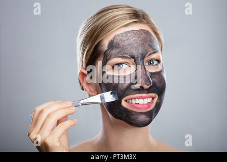 Jeune femme l'application de charbon activé, masque de visage à l'aide d'un pinceau sur fond gris Banque D'Images