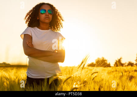 Belle course mixte African American female adolescent fille jeune femme portant sunglassesat coucher et au lever du soleil dans un champ de blé, de maïs ou d'orge Banque D'Images