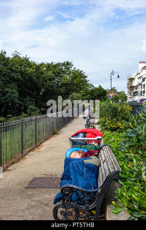 Tôt le matin, jeune femme sans-abri dorment dans la rue sur un banc de parc, ses possessions dans un seul panier. Banque D'Images