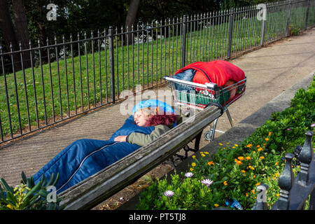 Tôt le matin, jeune femme sans-abri dorment dans la rue sur un banc de parc, ses possessions dans un seul panier. Banque D'Images