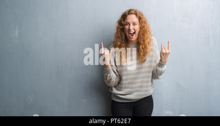 Jeune femme rousse sur mur gris grunge criant avec crazy expression faisant rock symbole avec les mains. Music star. Concept lourd. Banque D'Images