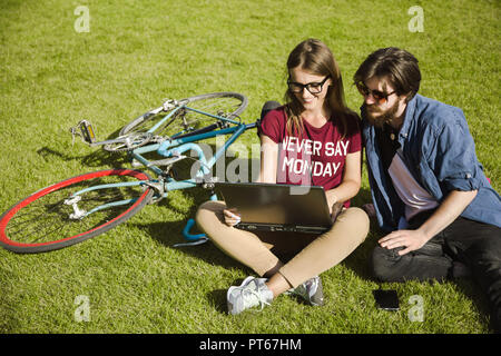 Les étudiants inspirés portant des lunettes assis sur l'herbe verte et de travail avec ordinateur portable, temps d'été ensoleillé Banque D'Images